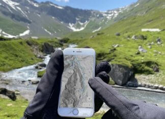Keine kalten Hände und Füße mehr beim Mountainbiken Hände mit warmen Handschuhen vor einer Bergkullisse die ein Mobiltelefon halten